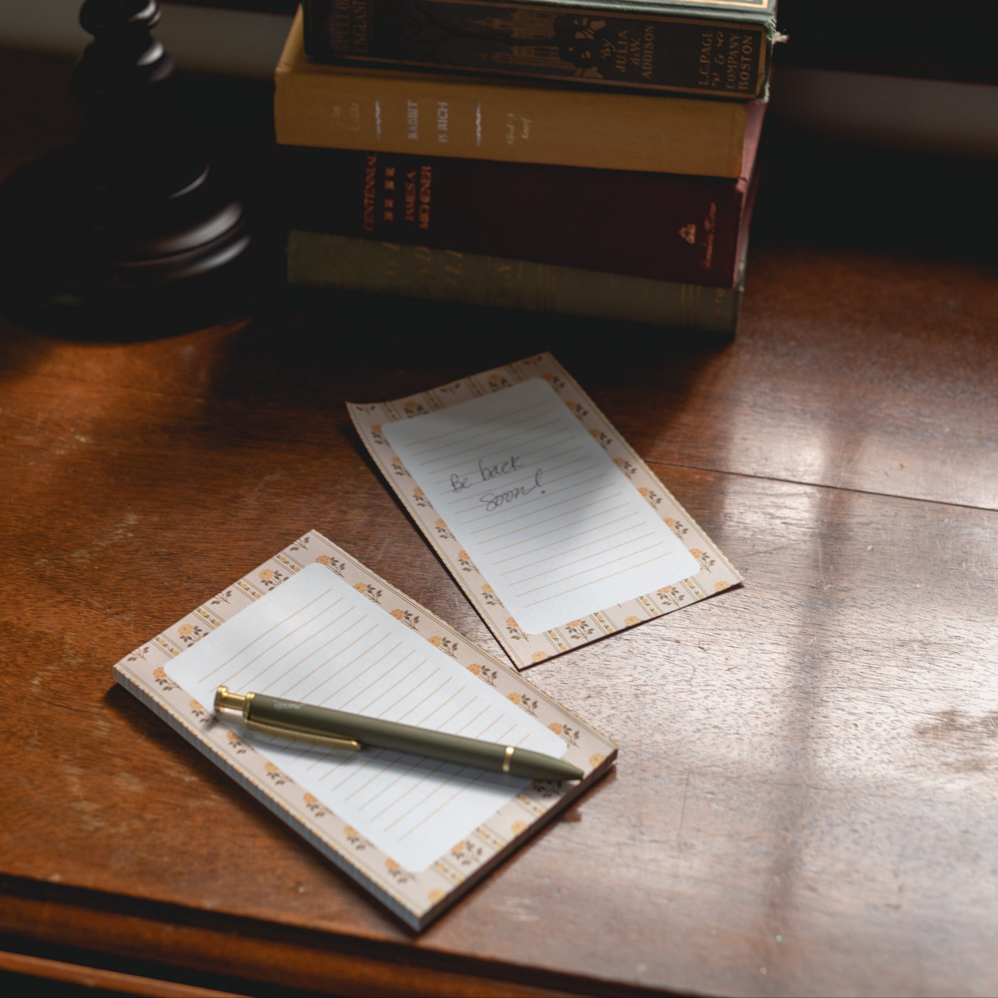 A stack of vintage books sits on a wooden windowsill next to a lamp, with two small lined notepads and a gold pen placed in front near an open window.