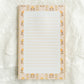 A lined notepad with a cream and yellow floral border, laid flat on a white lace fabric background.