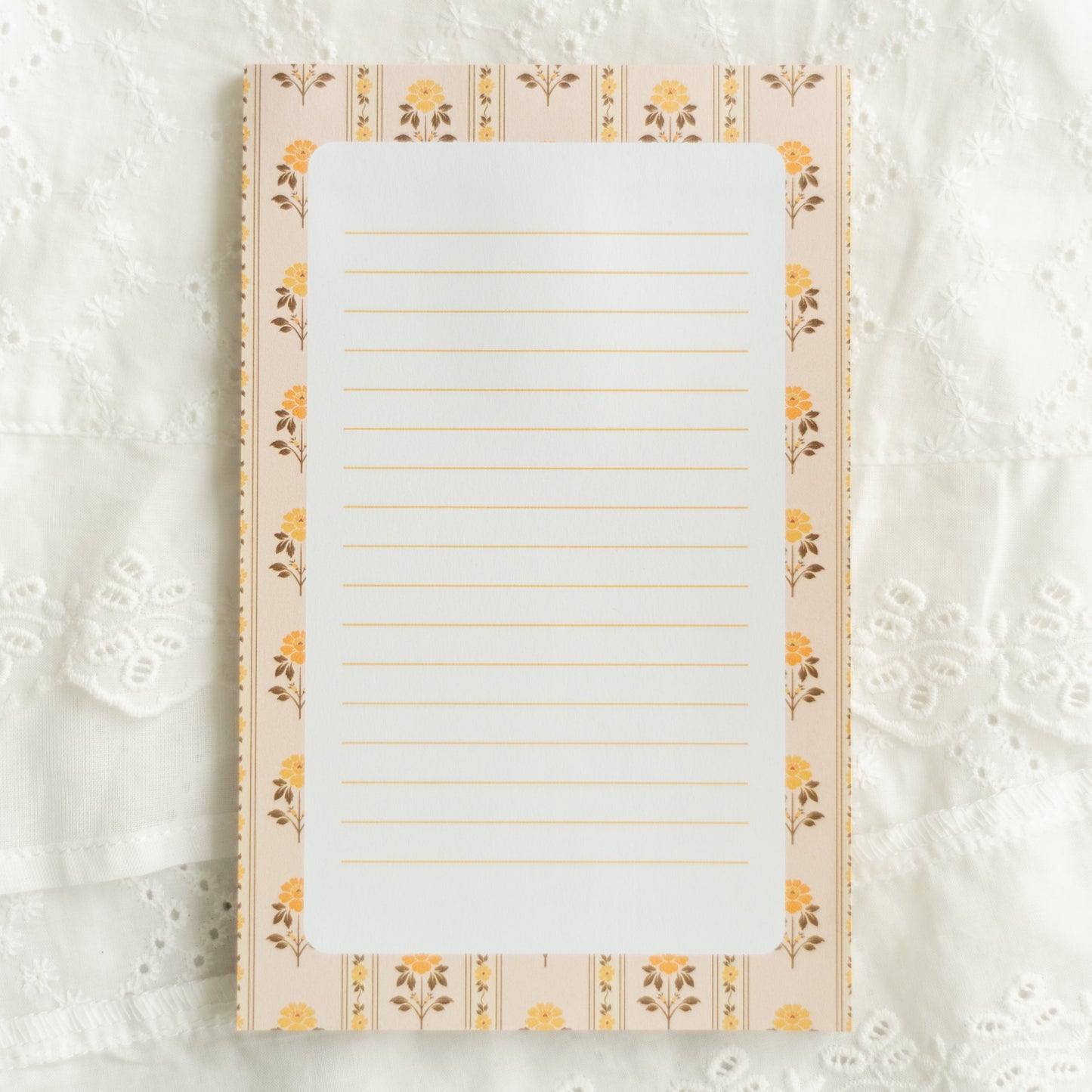 A lined notepad with a cream and yellow floral border, laid flat on a white lace fabric background.