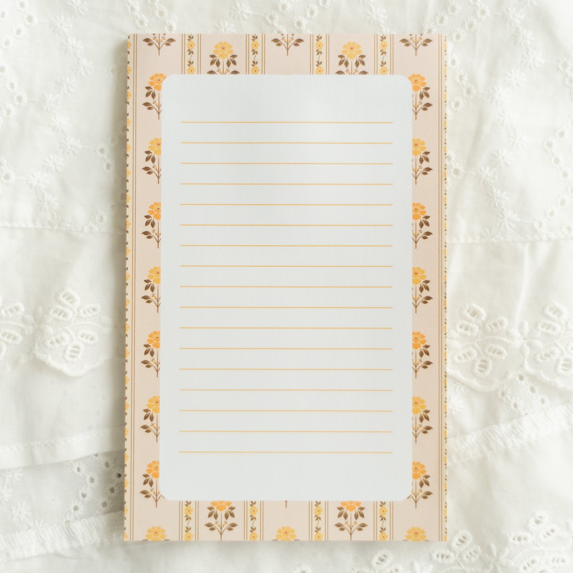 A lined notepad with a cream and yellow floral border, laid flat on a white lace fabric background.