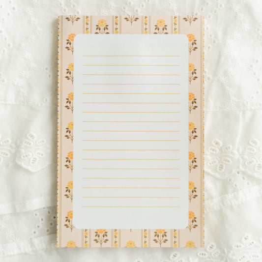 A lined notepad with a cream and yellow floral border, laid flat on a white lace fabric background.