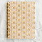 A spiral-bound notebook with a cream cover featuring a repeating yellow floral pattern, placed on a white lace fabric background.