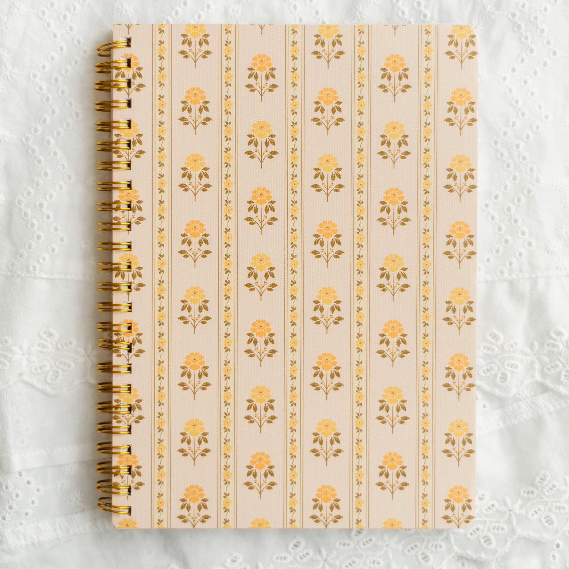 A spiral-bound notebook with a cream cover featuring a repeating yellow floral pattern, placed on a white lace fabric background.