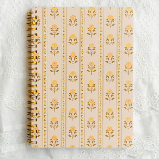 A spiral-bound notebook with a cream cover featuring a repeating yellow floral pattern, placed on a white lace fabric background.