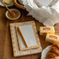 A styled tabletop scene with a floral notepad and matching pen in a wicker basket, alongside butter, honey, and a croissant on a wooden table.