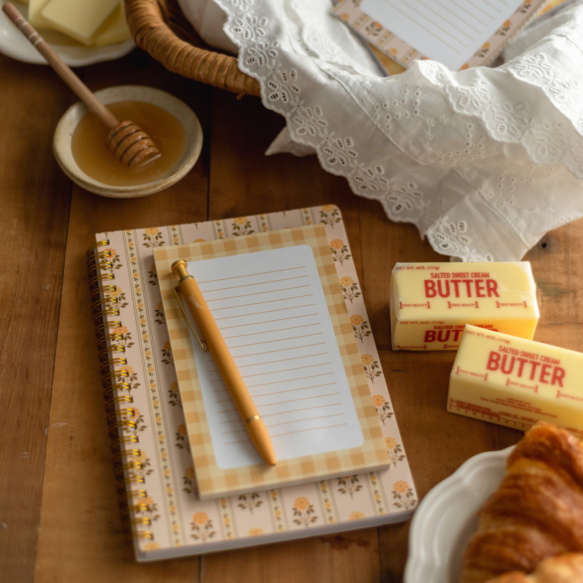 A styled tabletop scene with a floral notepad and matching pen in a wicker basket, alongside butter, honey, and a croissant on a wooden table.