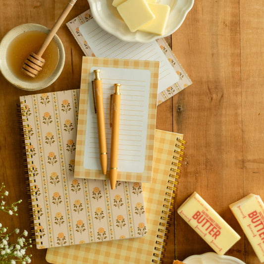 A styled flat-lay on a wooden table featuring patterned and gingham notebooks, two wooden pens, a small bowl of honey with a dipper, butter cubes on a plate, and butter sticks with pastry pieces.