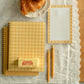 A styled flat-lay of a yellow and white gingham notepad set on a white lace table runner, featuring a spiral notebook, a lined notepad, two gold pens, a stick of butter, and croissants on a plate.