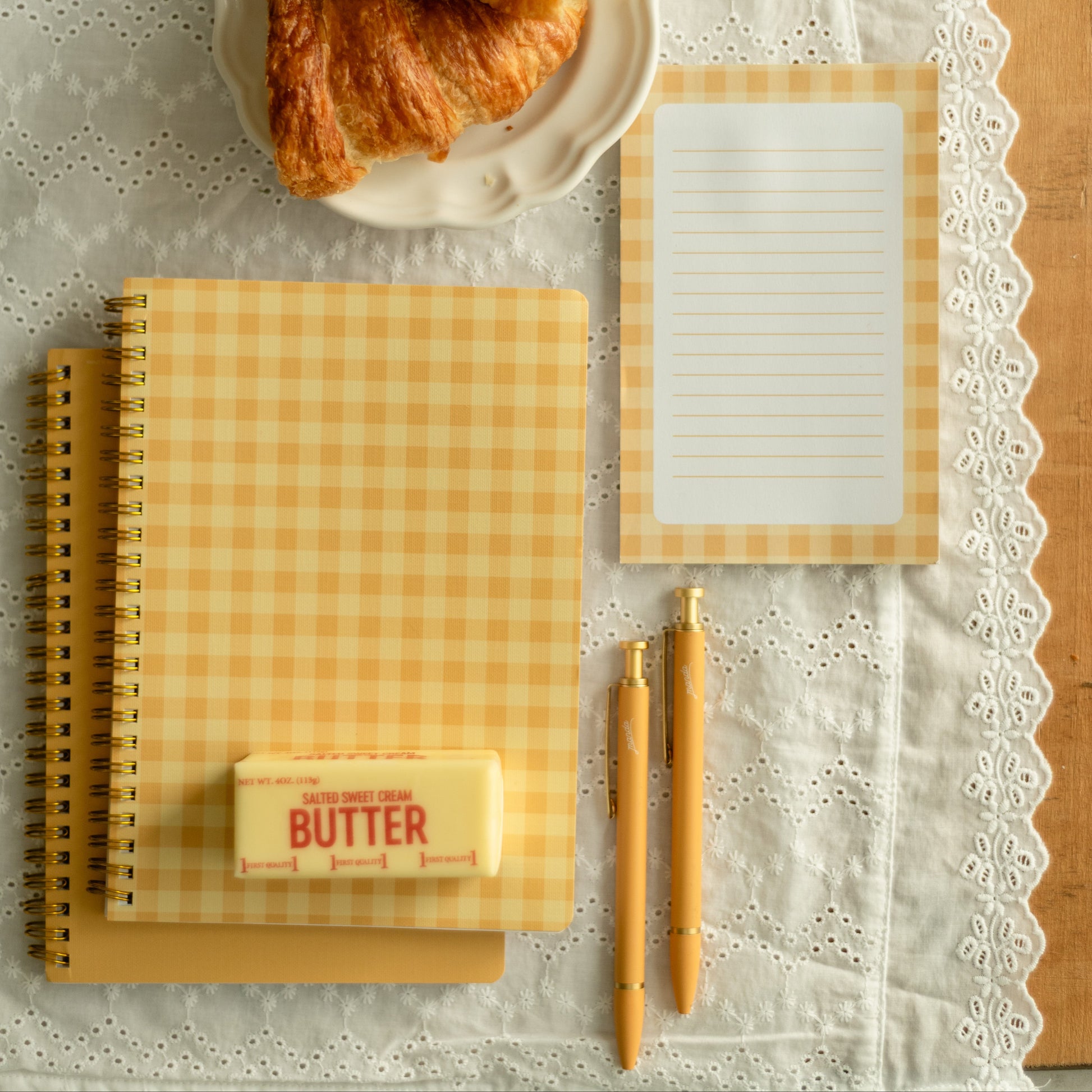 A styled flat-lay of a yellow and white gingham notepad set on a white lace table runner, featuring a spiral notebook, a lined notepad, two gold pens, a stick of butter, and croissants on a plate.