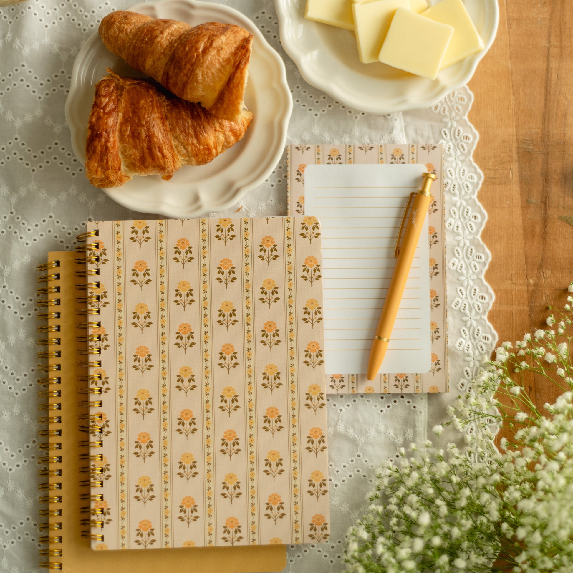 A styled flat-lay featuring a patterned notebook with a spiral binding, a lined notepad with a yellow pencil, and two croissants on plates with butter, arranged on a white lace cloth over a wooden table.