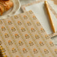A close-up of a spiral-bound notebook with a beige cover decorated with repeating yellow and green floral motifs. A matching gold pen rests on a lined paper sheet in the background, with a croissant partially visible on a plate in the upper left.
