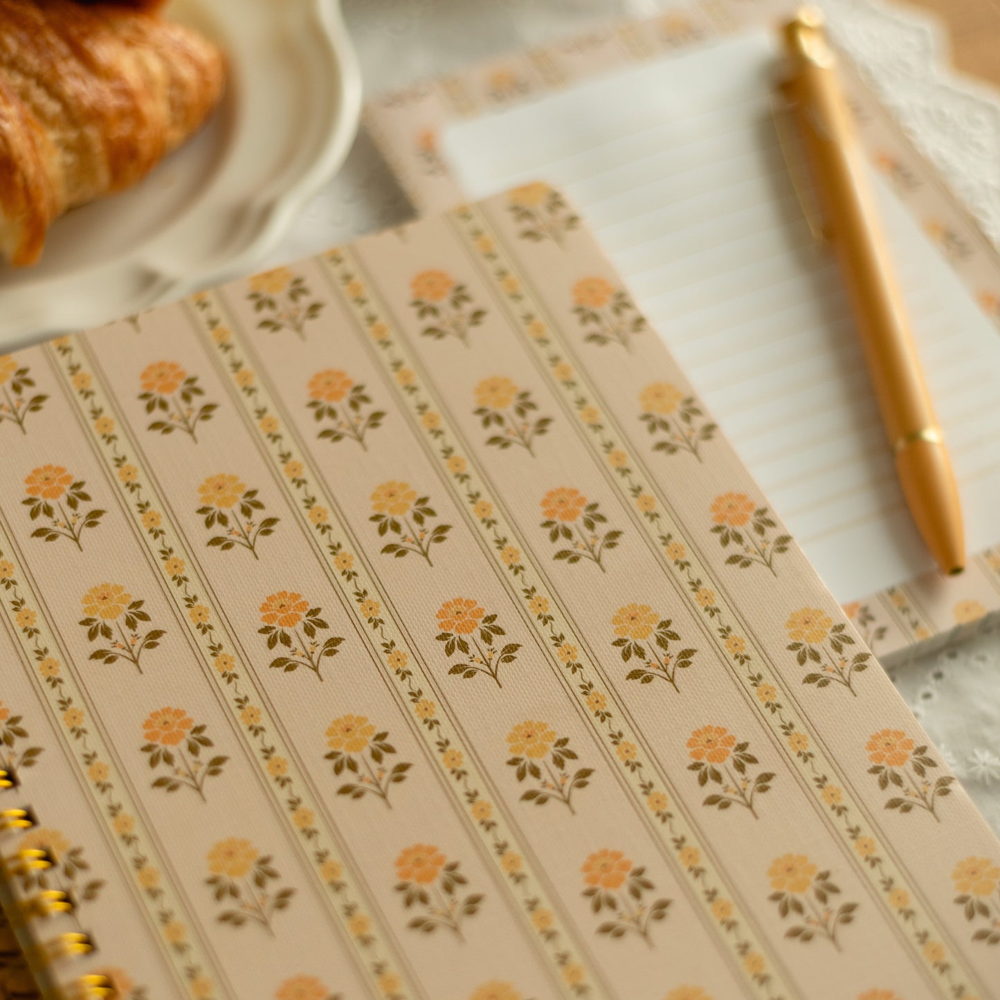 A close-up of a spiral-bound notebook with a beige cover decorated with repeating yellow and green floral motifs. A matching gold pen rests on a lined paper sheet in the background, with a croissant partially visible on a plate in the upper left.