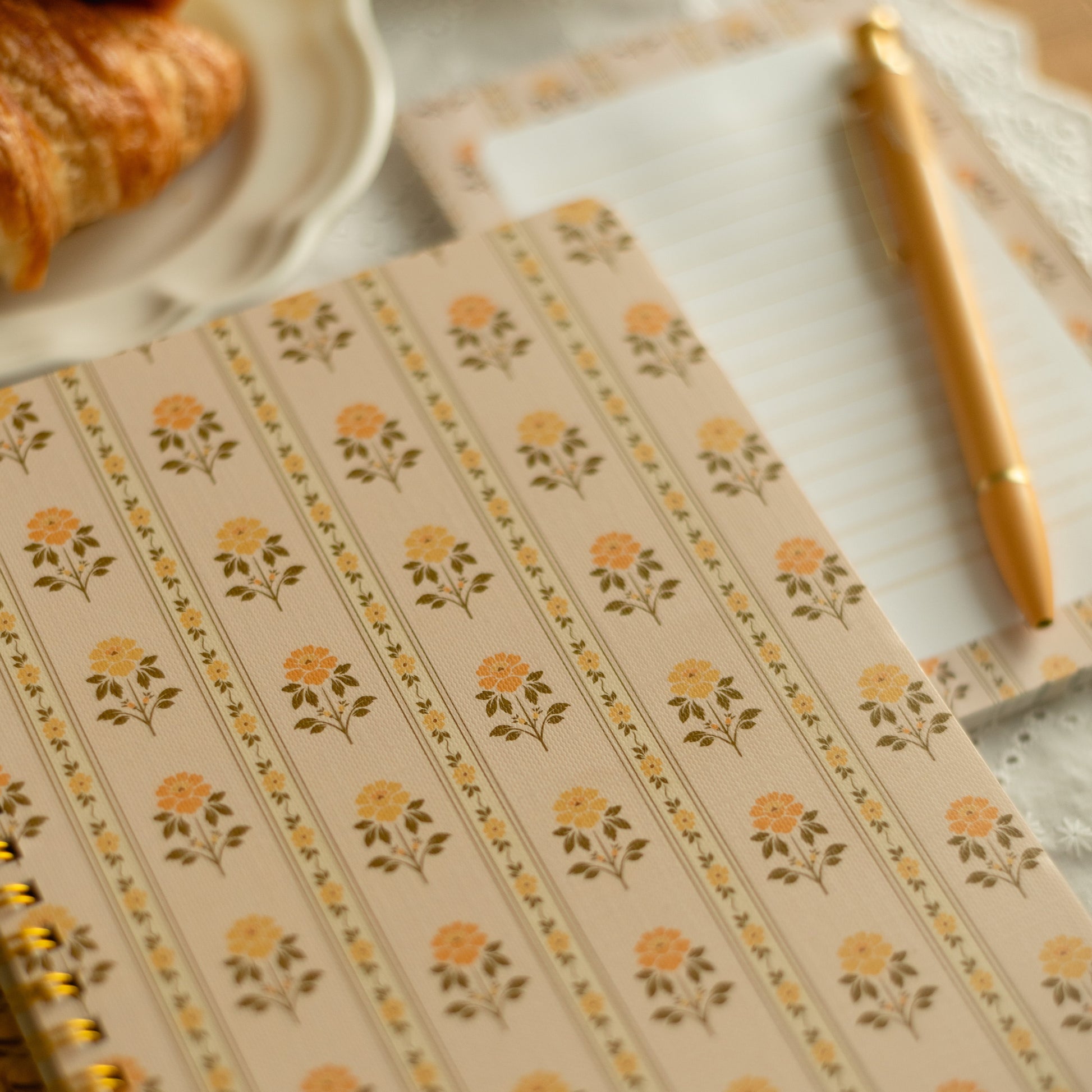 A close-up of a spiral-bound notebook with a beige cover decorated with repeating yellow and green floral motifs. A matching gold pen rests on a lined paper sheet in the background, with a croissant partially visible on a plate in the upper left.