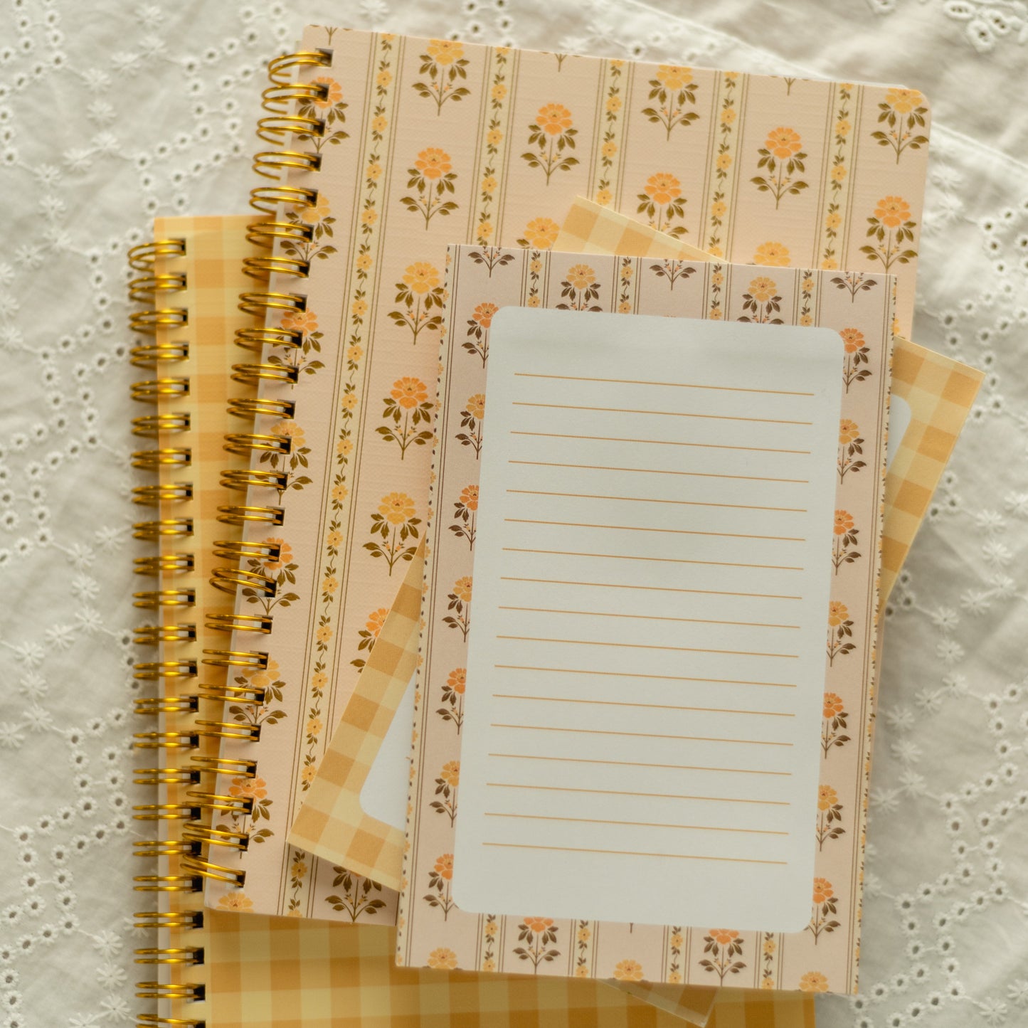 A stack of spiral-bound notebooks with floral and gingham patterns in soft yellow, peach, and cream tones, arranged on a white embroidered lace fabric background.