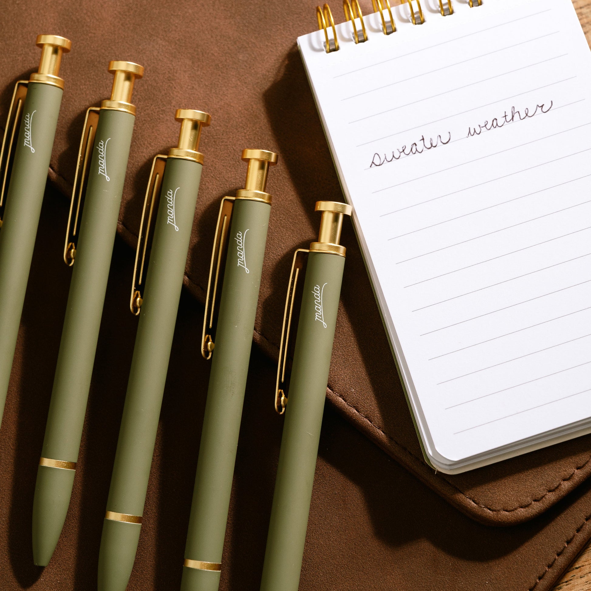 Set of green pens with gold accents on a brown leather surface next to a small notebook.