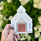 Hand holding a small house-shaped ornament in front of green foliage