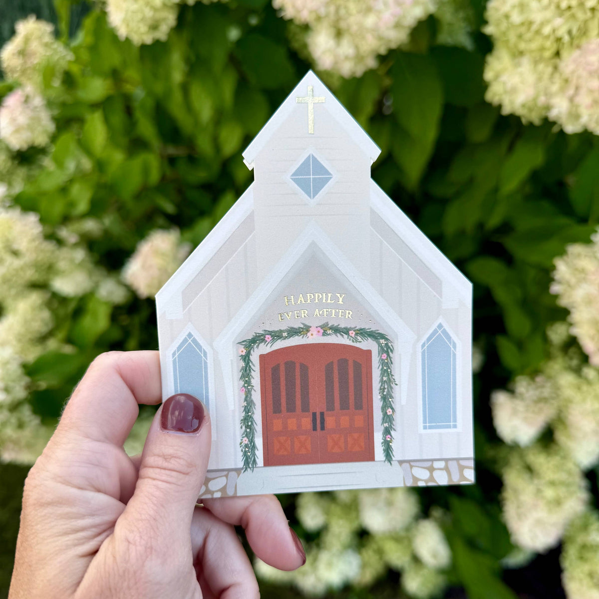 Hand holding a small house-shaped ornament in front of green foliage